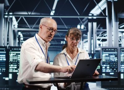 Senescent colleagues looking around high tech data center, using laptop to crosscheck disaster recovery plan and assess server machines in need of replacement, preventing glitches